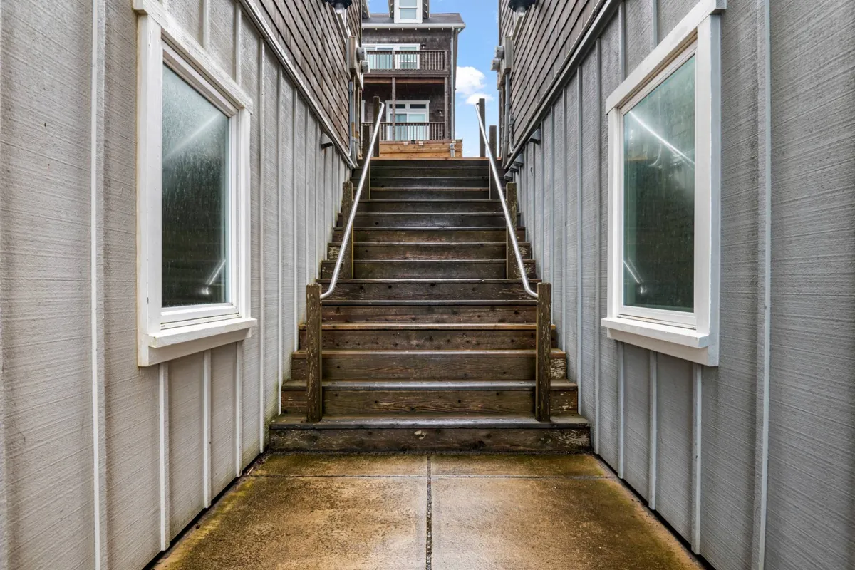 An inviting exterior shot showing wooden stairs leading up to the home, flanked by side walls and windows. The perspective creates a welcoming approach, suggesting the start of a relaxing stay for guests and their pets.