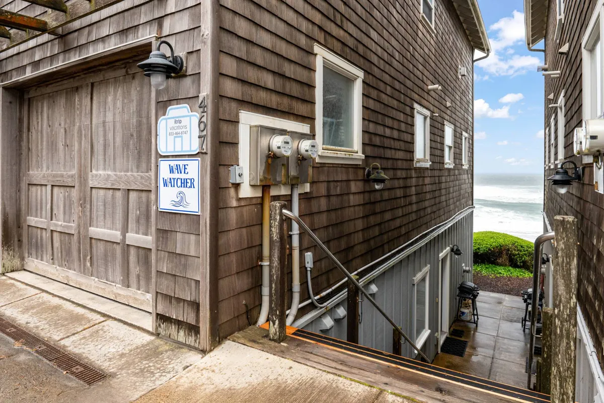 This image features the exterior of Wave Watcher, highlighting its wooden garage door, Wave Watcher sign, and a clear view of the ocean in the background. The image exudes a sense of direct access to the beach and the relaxed vibe of a coastal getaway.