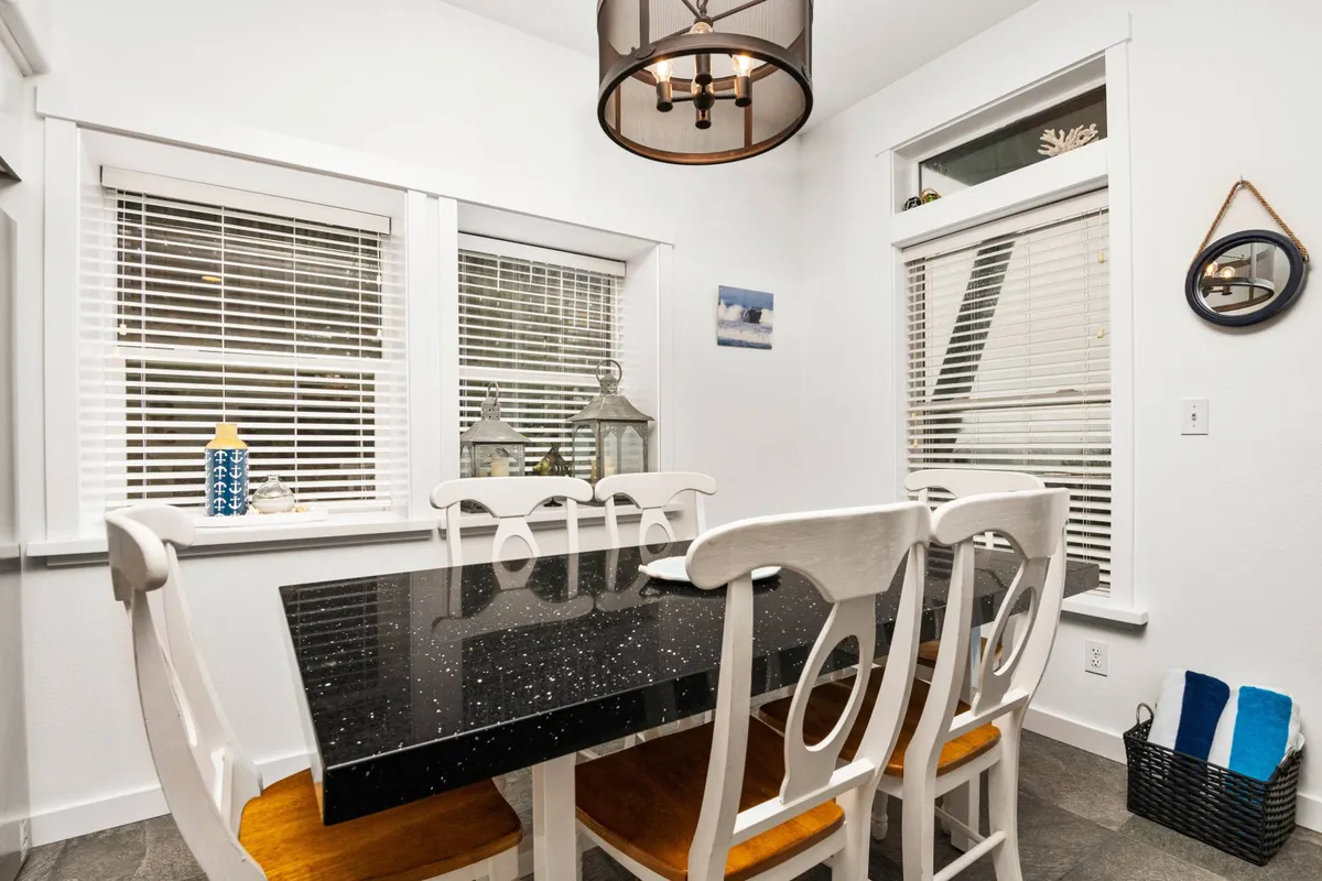 A cozy dining nook centered around a black granite dining table, surrounded by white wooden chairs and illuminated by a stylish chandelier. Decorative items adorn the windowsill, and the space is awash in natural light, setting a warm and inviting atmosphere for shared meals.