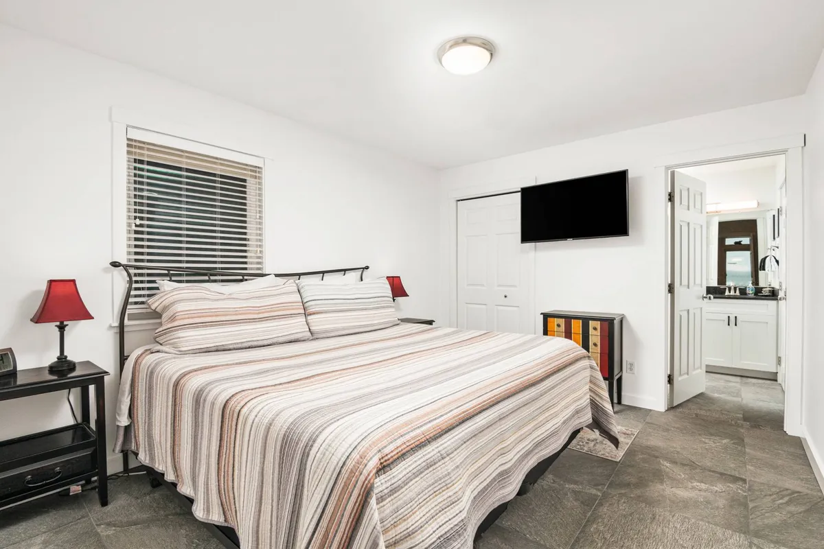 A cozy bedroom featuring striped bedding on a king-size bed, complemented by red lamps on the nightstand and a wall-mounted TV. The doorway to the en-suite bathroom is also visible, providing a glimpse of the suite's layout and convenience.