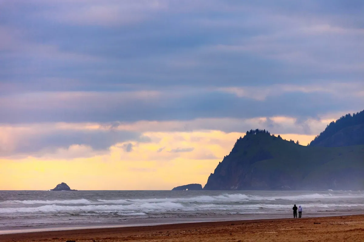 A romantic scene featuring a couple walking hand-in-hand along a sandy beach, with gentle waves lapping at the shore and distant cliffs providing a dramatic natural backdrop. The lighting suggests either an early morning or sunset, enhancing the mood of intimacy and tranquility.