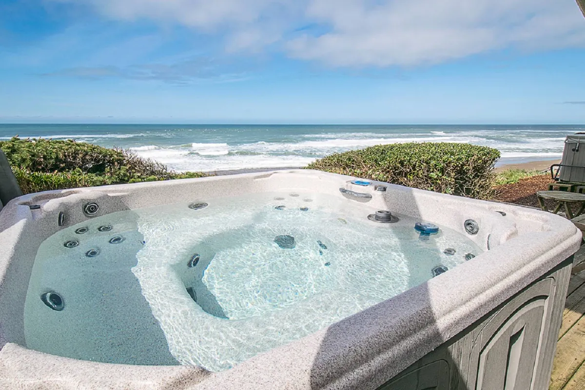A private deck with a bubbling hot tub overlooks the Pacific Ocean, framed by lush landscape and open sky. The scene exudes tranquility, inviting guests to relax in the hot tub while enjoying starlit nights or stunning sunsets over the water.