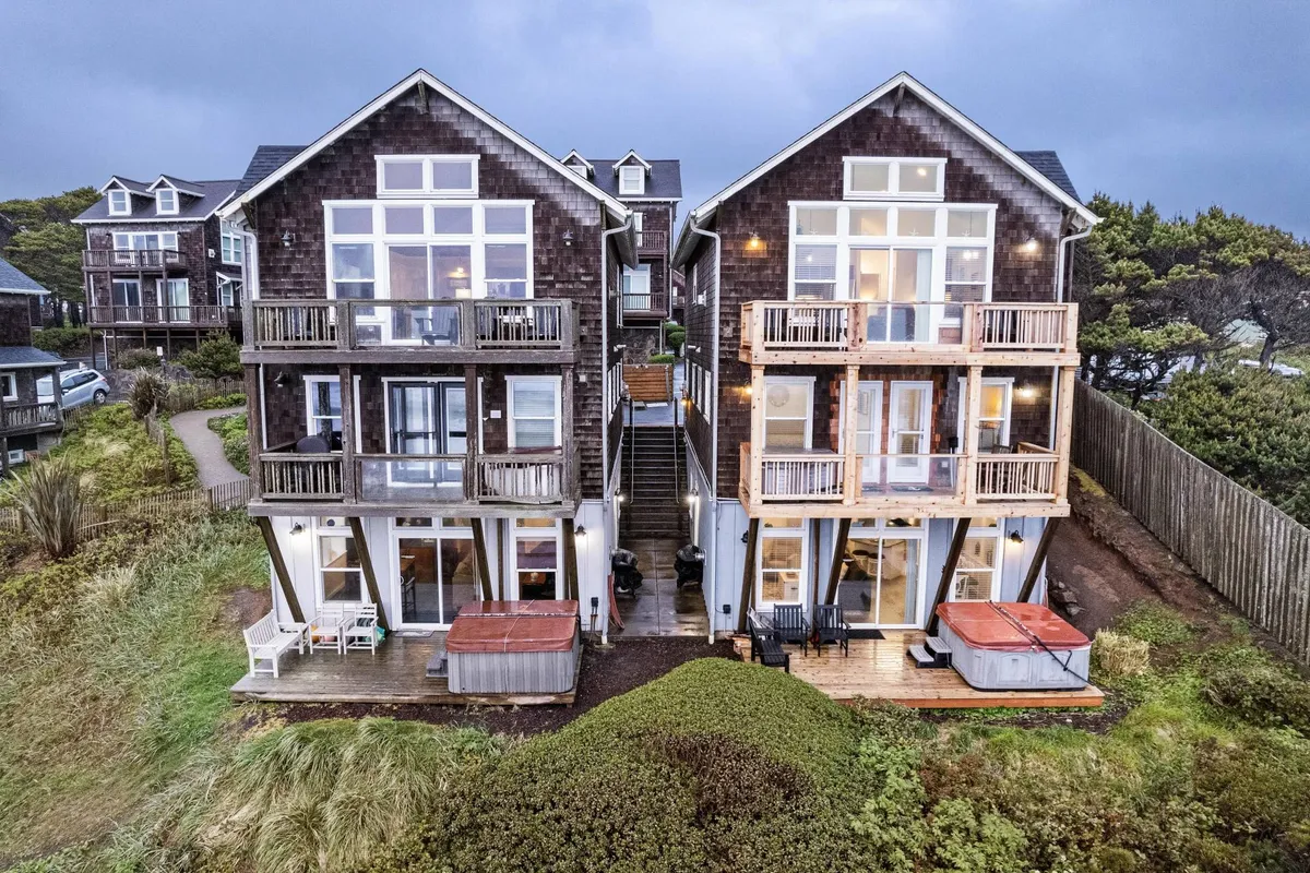 This exterior image features two large homes with inviting balconies and visible hot tubs, connected by a staircase. People are seen relaxing in the hot tubs and enjoying social time on the balconies, with the ocean view in the background. The scene is cozy and welcoming, suggesting a setting where groups can gather and unwind together.