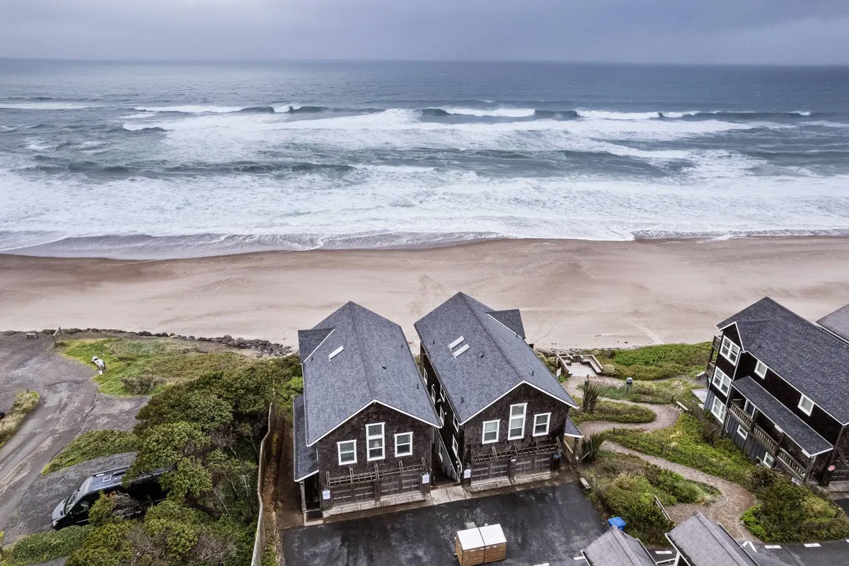 A panoramic exterior view of beachfront houses nestled behind sand dunes, with rolling ocean waves and a calm, expansive shoreline in the foreground. The scene is peaceful and invites thoughts of morning strolls and sunset watching.
