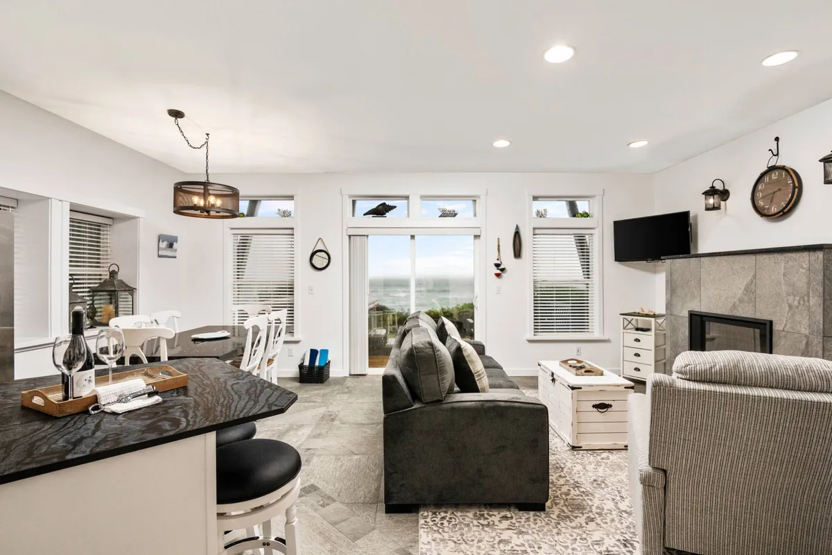 The image showcases a bright, inviting living room with plush seating positioned to take in sweeping ocean views through expansive windows. A stylish bar counter with stools offers a spot for morning coffee or evening cocktails, while warm touches like a decorative clock and modern light fixtures add coziness. The Pacific Ocean is visible just beyond, and the space is bathed in natural light ideal for relaxing, sharing meals, or simply enjoying a sunset.