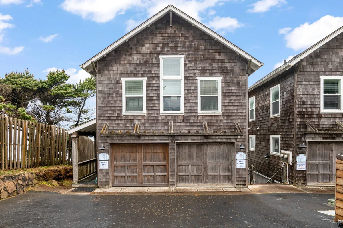 A welcoming exterior view of a home with classic wooden shingles, large windows, and a garage door, surrounded by lush greenery. The pathway leading to the entrance invites strolls, and the environment radiates peace and community charm.